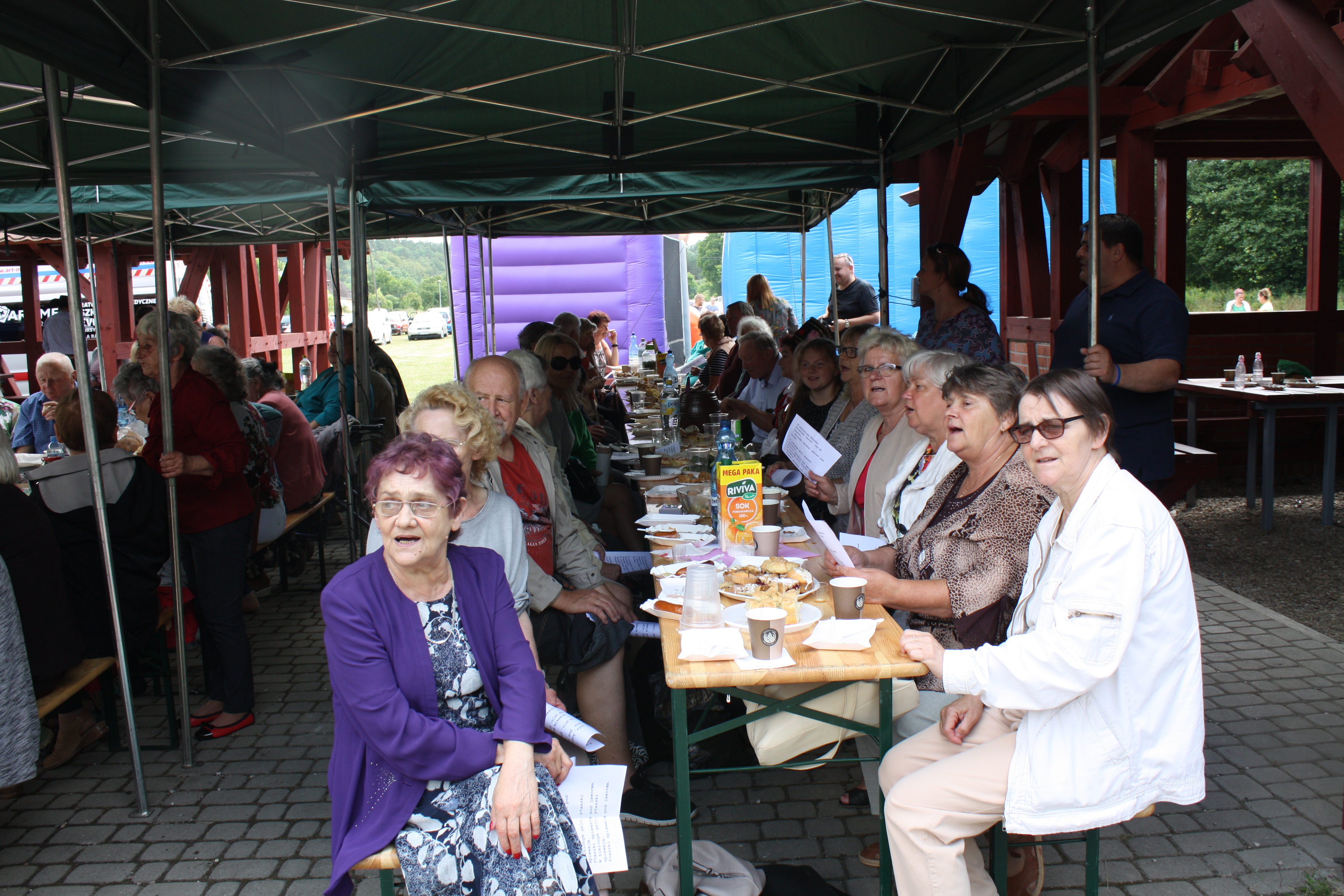 Uczestnicy spotkania siedzący przy biesiadnych stołach pod wiatą, podczas wspólnego śpiewu. 