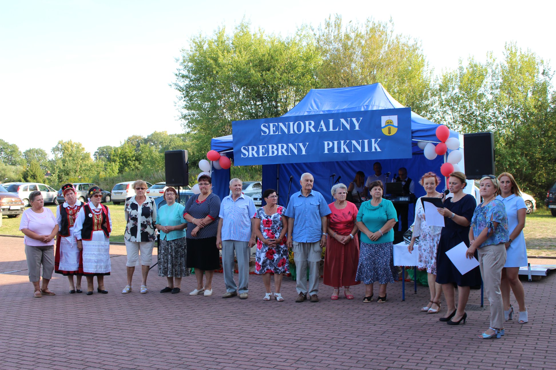 Grupa seniorów zgłoszonych do konkursu "Srebrne Serce" na tle namiotu - sceny Senioralnego Srebrnego Pikniku.