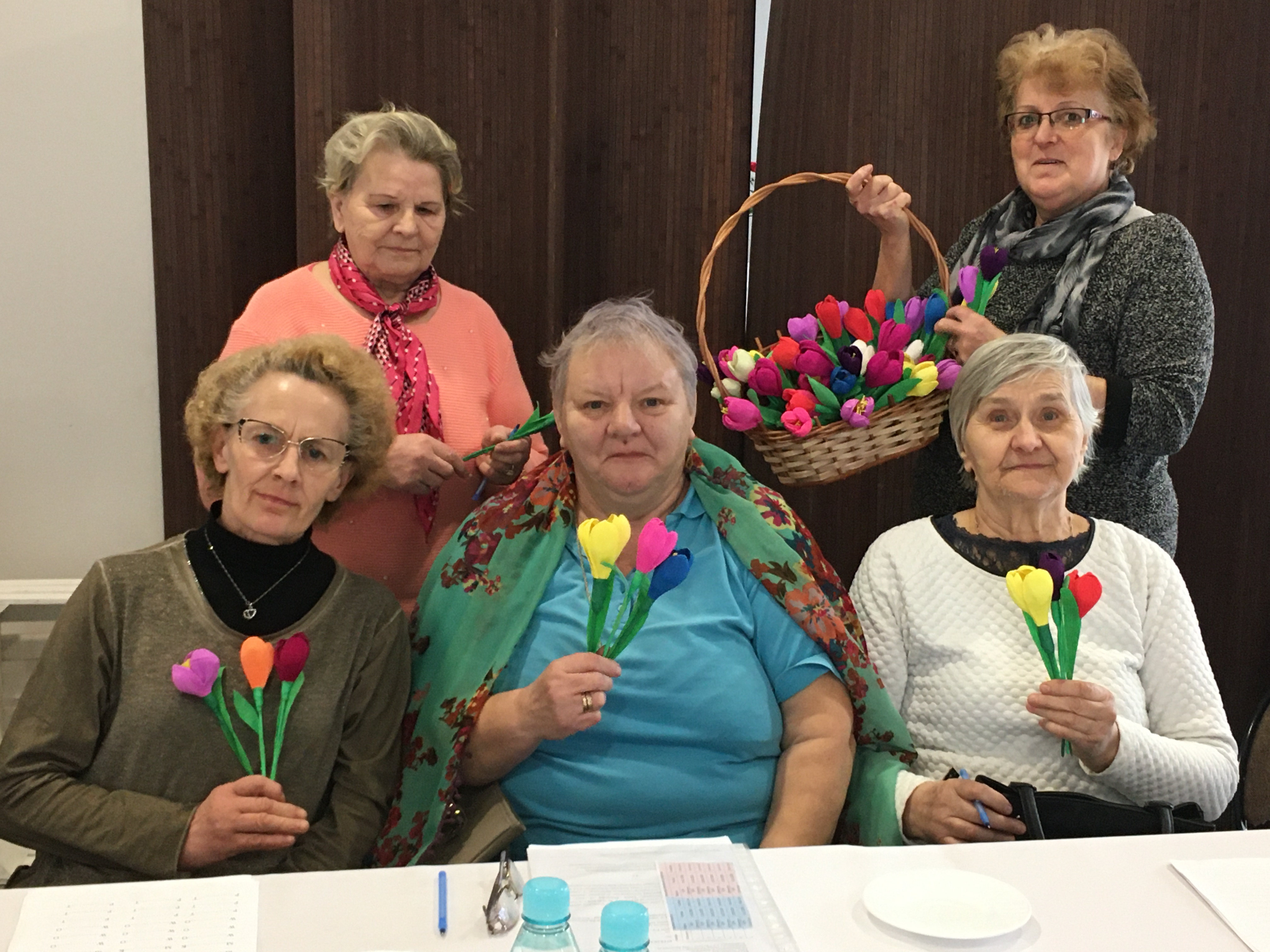 Seniorki siedzące przy stole trzymające w ręce bukiety otrzymanych kwiatów z okazji dnia kobiet.