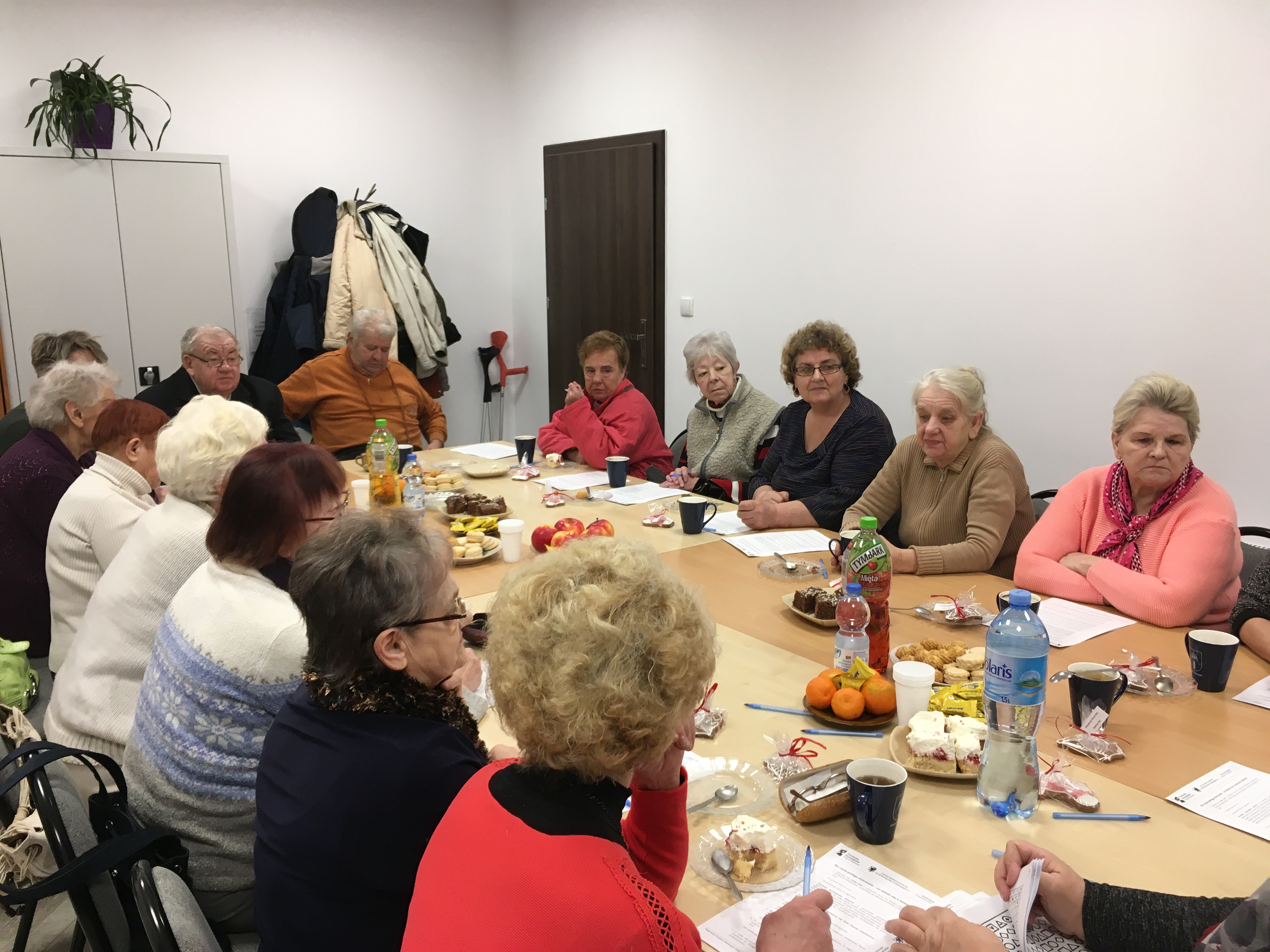 Seniorzy skupieni wokół stołu wsłuchujący się w prowadzące szkolenie trenerki.